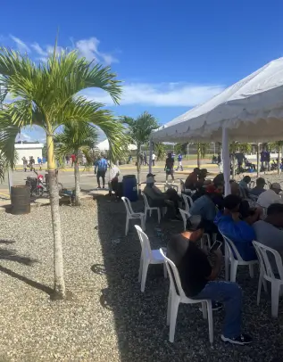 Spectators in plastic chairs watch the baseball game under a large tent